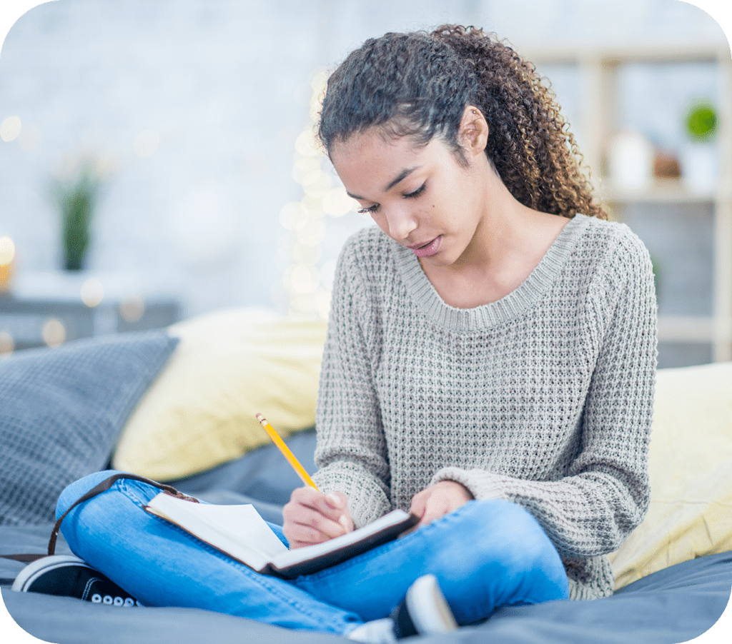 A young person sitting on their bed and writing in a notebook.