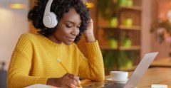 Smiling black girl with wireless headset studying online, using laptop at cafe, taking notes, copy space